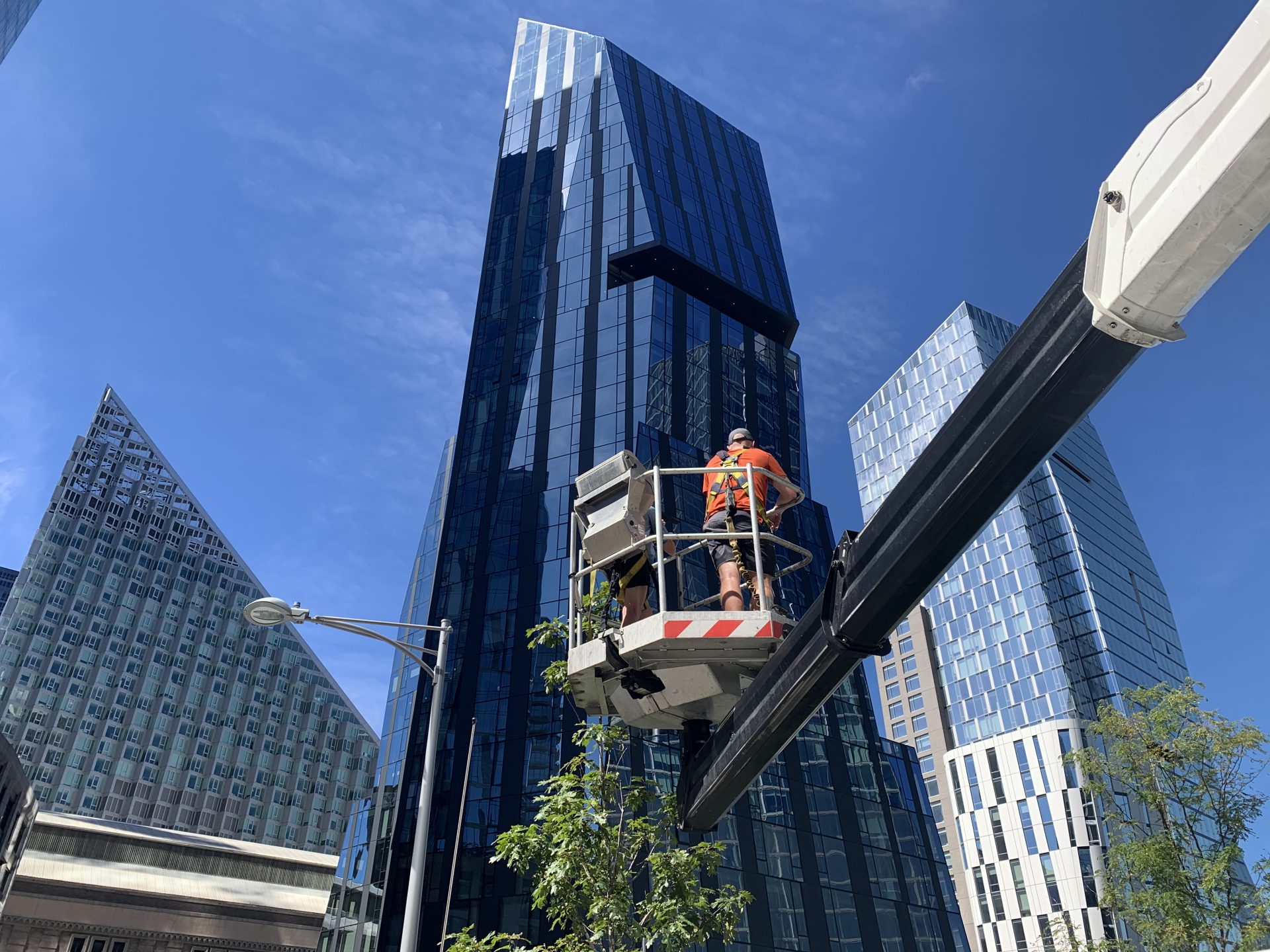 Two Skyway technicians in safety harnesses in an aerial lift basket, rising past glass towers under a blue sky