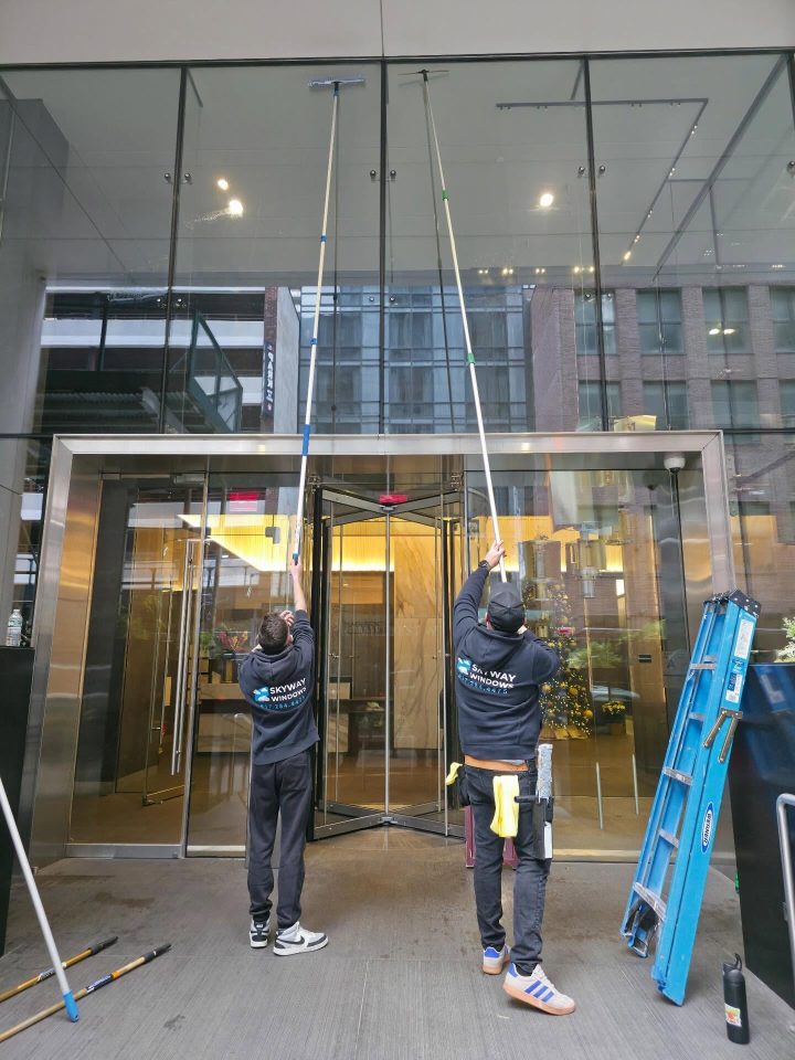 Two Skyway technicians cleaning the glass entrance of a new building with water-fed poles